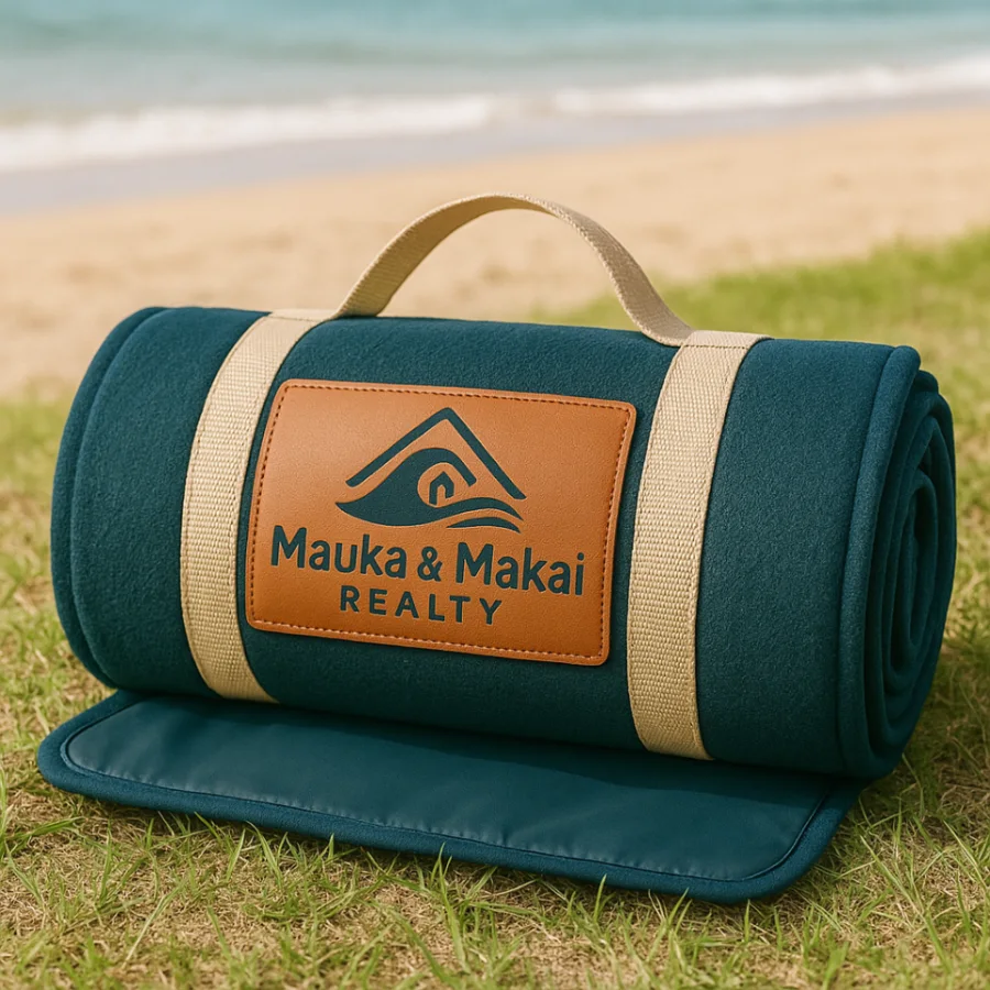 A rolled teal beach blanket with a tan leather patch carrying the Mauka and Makai Realty logo, on grass above a beach.