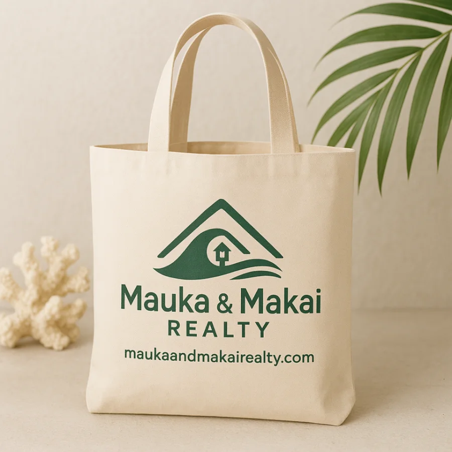 A natural canvas tote bag beside coral and a palm frond, printed in green with the Mauka and Makai Realty mark, wordmark and web address.