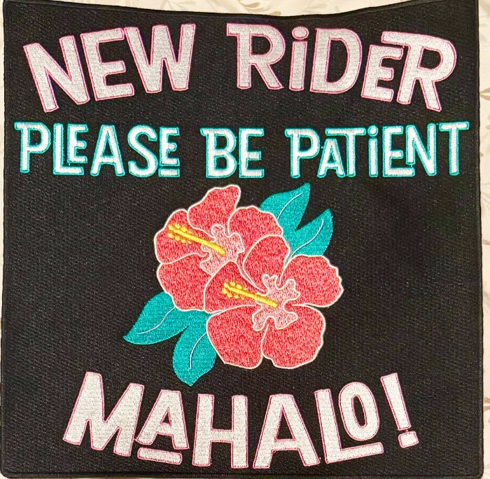 Photograph of the finished embroidered back patch: New Rider, Please Be Patient, Mahalo! stitched in white, pink and teal around a red hibiscus on black twill.